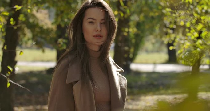 Charming Caucasian girl with pleasant smile standing behind yellow leaves in park and shaking her long brown hair. Pretty European woman posing at the camera outdoors. Cinema 4k footage ProRes HQ.