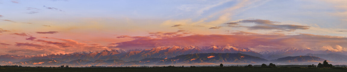 Panorama. Fairytale dawn, sunset with bright multi-colored clouds, against the backdrop of a mountain range with snow-capped peaks