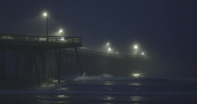 Wide view of Pismo Beach Pier at night, with lights reflected in the water and fog rolling in over the Pacific Ocean.