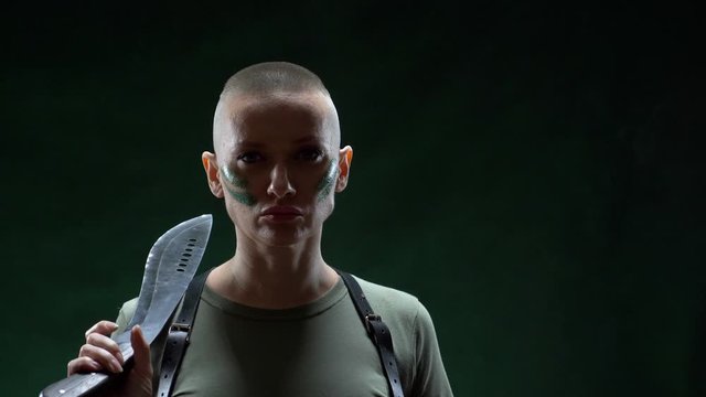 Angry bald girl with camouflage makeup shaving machete hair on a black background