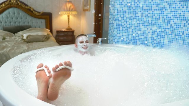 a bald young woman with a mask on her face lies in a bath with foam, her legs with toe separator are visible from the water.