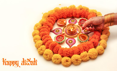 Diwali background with woman hand lighting clay lamps with traditional rangoli decorated with marigold flowers on white background