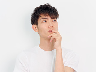 Fototapeta na wymiar Portrait of a handsome Chinese young man in white t shirt looking away with hand on his lips, thinking expression, isolated on white background. 