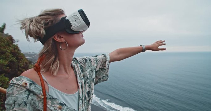 Young modern woman entertaining with VR-headset enjoying spectacular scenery of ocean and volcanic coastline of Tenerife island. Freedom, traveling and technology concept.