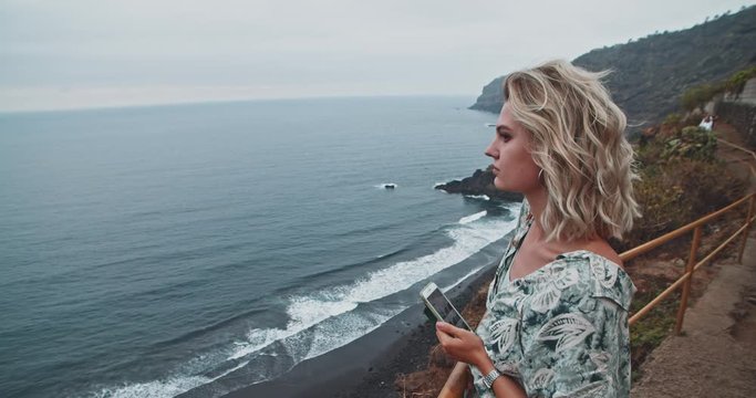Beautiful young woman overlooking glorious calm ocean on cliffside viewpoint. Delighted tourist girl taking pictures of amazing nature scenery in Tenerife.