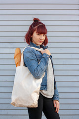 Long french bread in an eco textile bag on the shoulder of a young girl with red hair. Girl in a denim jacket and black pants.