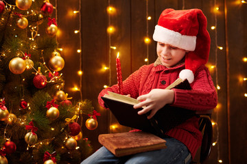 Obraz na płótnie Canvas Teen boy reading book, sitting indoor near decorated xmas tree with lights, dressed as Santa helper - Merry Christmas and Happy Holidays!