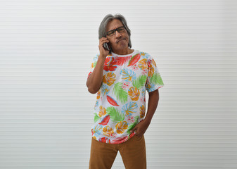 Thoughtful elderly traveler asian man wearing summer shirt and glasses talking with smart mobile phone over white wall, Funny face expression pose, Business summer holiday concept