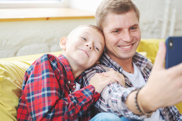 Obraz premium Young father with a son, a boy of 8 years old in checkered shirts, sitting on a yellow sofa, taking a selfie on a mobile phone, time together, relationship, family, one parent concept