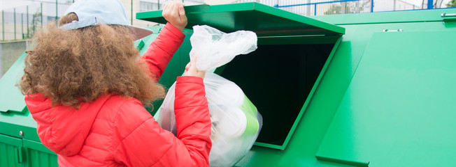a girl in a red jacket, throws a full package into a green dumpster