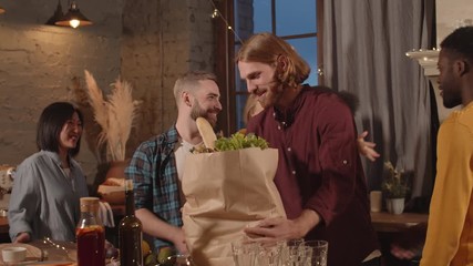 Tracking shot of multi-ethnic group of happy friends having dinner party in cozy apartment. Home owners smiling and hugging guests arriving with groceries