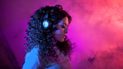 Portrait of a curly pensive sensual woman in big headphones, with a soft smile listening to music, on neon background.