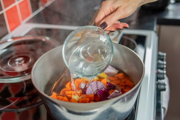 Selective focus of a home cooking chef pouring stock in to a saucepan of chopped carrots, ginger, red onions and garlic and making a healthy homecooked soup