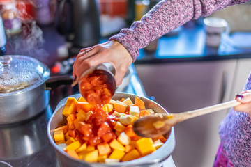 Selective focus of a home cook adding a can of chopped tomatoes to a stainless-steel pan root vegetables to create a nutritious vegetarian paprika stew
