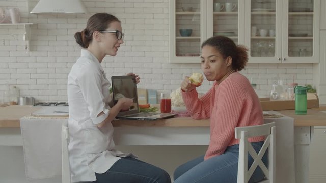 Nutritionist showing personalized diet plan on laptop for apathetic african woman during appointment in domestic kitchen. Indifferent overweight woman ignoring recommendation of dietitian at home.