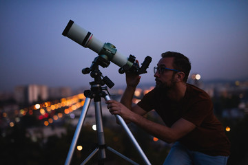 Astronomer with a telescope watching at the stars and Moon with blurred city lights in the background.