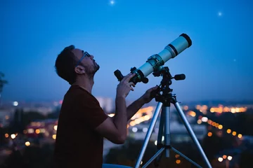 Fotobehang Astronomer with a telescope watching at the stars and Moon with blurred city lights in the background. © astrosystem