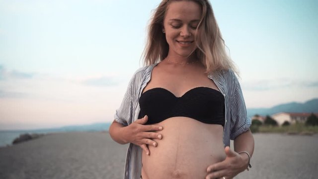 Attractive young pregnant woman and her tiny belly with unborn baby. Happy woman embracing lovely her tummy and smiling. Expectant woman dancing at the beach in sunset. Maternity concept