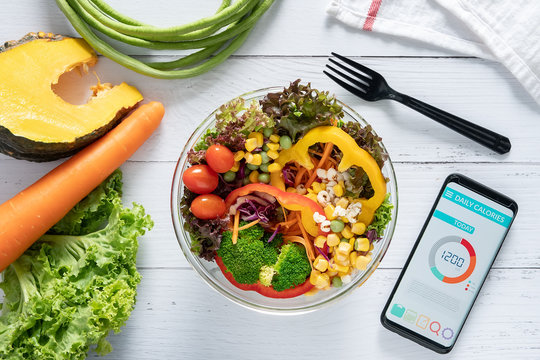 Calories counting , diet , food control and weight loss concept. Calorie counter application on smartphone screen at dining table with salad, fruit juice, bread and vegetable