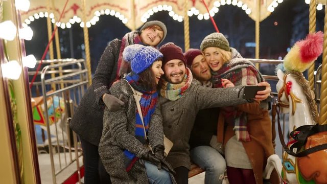Happy people taking selfie photo on smartphone camera at New Year winter fair amusement park. Smiling friends have fun riding carousel at Christmas market. Blogger works. Garland lights on backdrop.