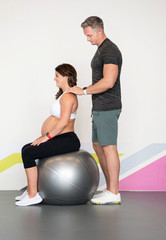 Caucasian pregnant woman in her third trimester with her personal trainer doing bodyweight exercises on a rubber ball  wearing sports or fitness clothing