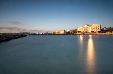 Fototapeta na wymiar Cala Bona looking towards Cala Millor and the Protur Alicia Hotel at sunrise. Beautiful reflections on the sun in the water which is calm and silky smooth.