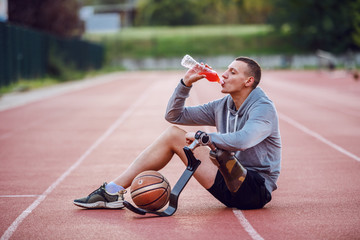 Handsome caucasian sporty handicapped man in sportswear sitting on racetrack and drinking refreshment. Between legs is basketball ball.