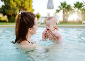 Fototapeta na wymiar Cheerful Caucasian 6 months old baby boy learning how to swim at swimming pool. Mother holding her son. First time at pool concept.
