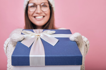 A cheerful happy girl stands with a pink back wall in glasses, milk mittens and hat, reaches out...