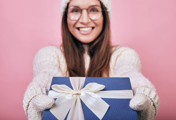 A cheerful happy girl stands with a pink back wall in glasses, milk mittens and a hat, reaches out with gift to you. Take a gift and give it to friends or relatives. Cool give joy. Smiling, glasses.