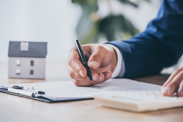 cropped view of businessman using calculator and writing in contract near house model