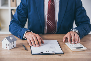 cropped view of businessman in formal wear using calculator and writing in contract near house model