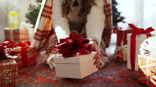 Close-up of christmas gift box with glittering red ribbon under the christmas tree. Girldressed in winter pyjamas with warm blanket sitting under the tree and opening her christmas present. Emotions