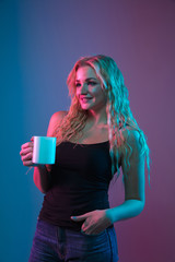 Caucasian young woman's portrait on gradient background in neon light. Beautiful female model with unusual look. Concept of human emotions, facial expression, sales, ad. Drinking coffee or tea.