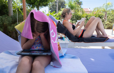 small child under a towel in the sun watching an iPad with her mother in the background reading a...