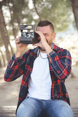Hombre joven con camisa de cuadros haciendo fotos con una camara instantanea en el bosque.