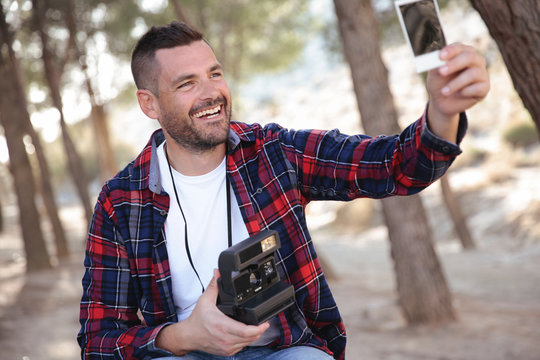 Hombre joven con camisa de cuadros obeservando una fotografia de una camara instantanea en el bosque.