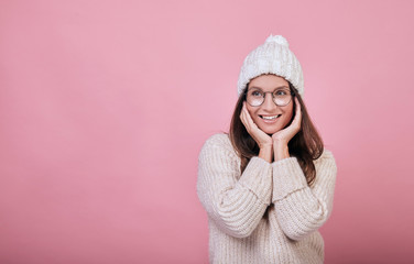 Cheerful smiling girl with an excellent face costs on a pink background. lady is wearing warm knitted sweater and a milk-colored hat, also in glasses. Warm clothes in cold season. Cosiness, comfort.