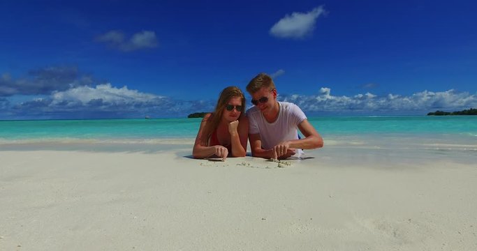Couple on vacation drawing a heart in the beautiful white sand beach of Australia on a sunny day with waves blue sky and clouds background - Steady shot