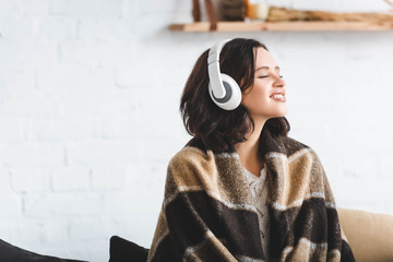 happy woman with closed eyes in blanket listening music with headphones - obrazy, fototapety, plakaty