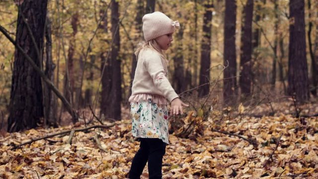 Happy young girl in pink hat plays in fall leaves in golden autumn forest