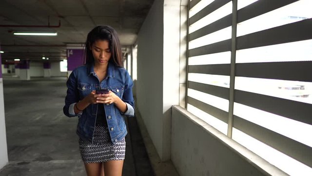 Attractive Asian teen texting while she walks through a parking garage and stops at a bright window