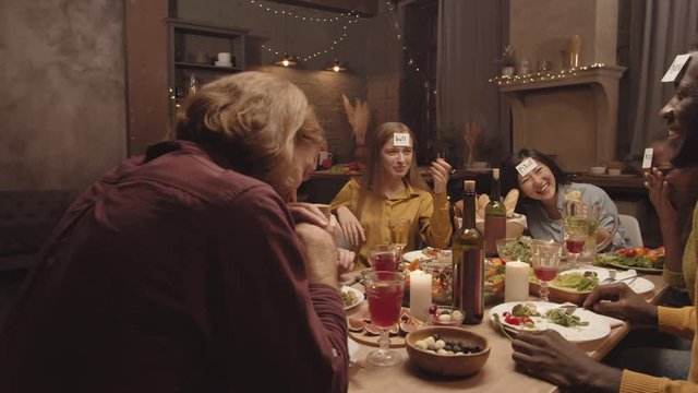 Handheld tracking of group of dinner guests playing &laquo;Who am I&raquo; game. They are laughing and answering questions of male friend trying to guess what is written on sticky note attached to his forehead