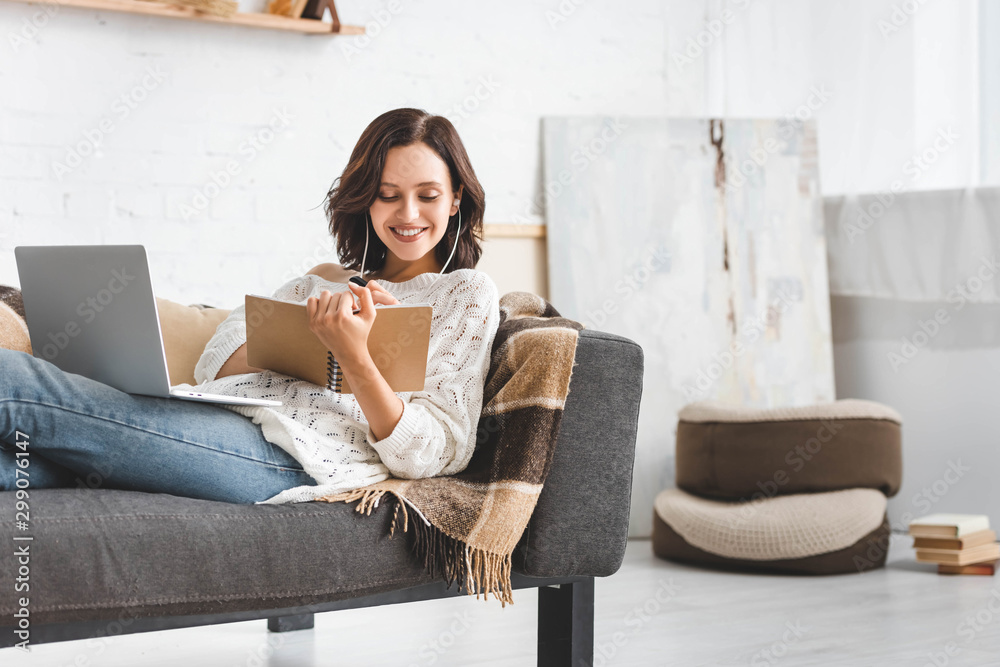 Wall mural attractive cheerful girl studying online with notepad, earphones and laptop