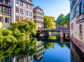 The Pont du Faisan spans the canal of the river Ill in the Petite France quarter in Strasbourg, France, lined with half-timbered houses reflecting in the still waters on a sunny morning. - obrazy, fototapety, plakaty