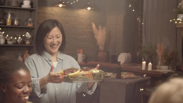 Tracking shot of happy Asian woman carrying delicious dish and putting it down on dinner table, then toasting and clanking glasses with cheerful group of friends