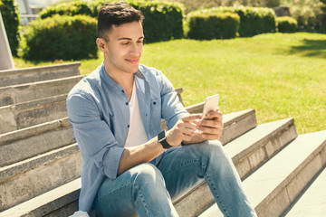 Fototapeta premium Outdoors Leisure. Stylish guy wearing watch sitting on stairs in park browsing smartphone smiling happy