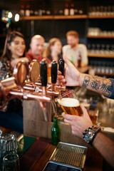 Cropped picture of tattooed barman pouring beer in glass. In background is group of friends sitting at counter. Pub interior.
