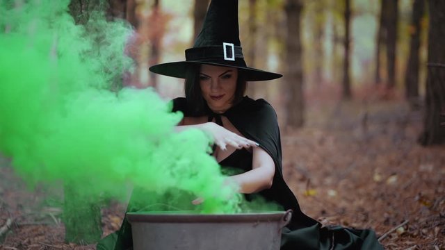  Black witch sits in front of cauldron from which green smoke is falling. Sorceress conjures, brews potion with spells. Horror, halloween, cosplay holiday, magic concept