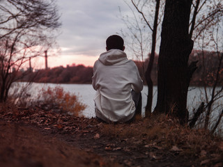 A lonely young man sits on the Bank of a river on a cold autumn evening copy space